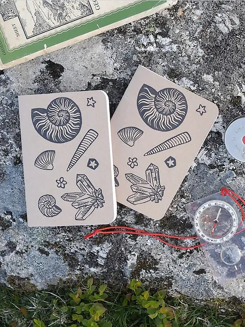 Zwei Notizbücher mit Linolschnitt-Einbänden aus Fossilien auf Stein mit Kompass und Karte von Becca Thorne