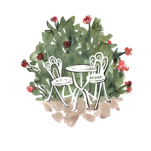White bistro table and two chairs set amidst green bushes with red flowers by Briana Kranz