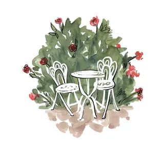 White bistro table and two chairs set amidst green bushes with red flowers by Briana Kranz
