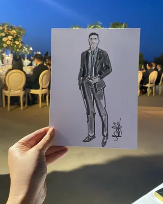 Hand holding sketch of man in black suit at outdoor gathering