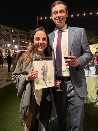 Smiling couple outdoors at night, holding portrait and wine glass.
