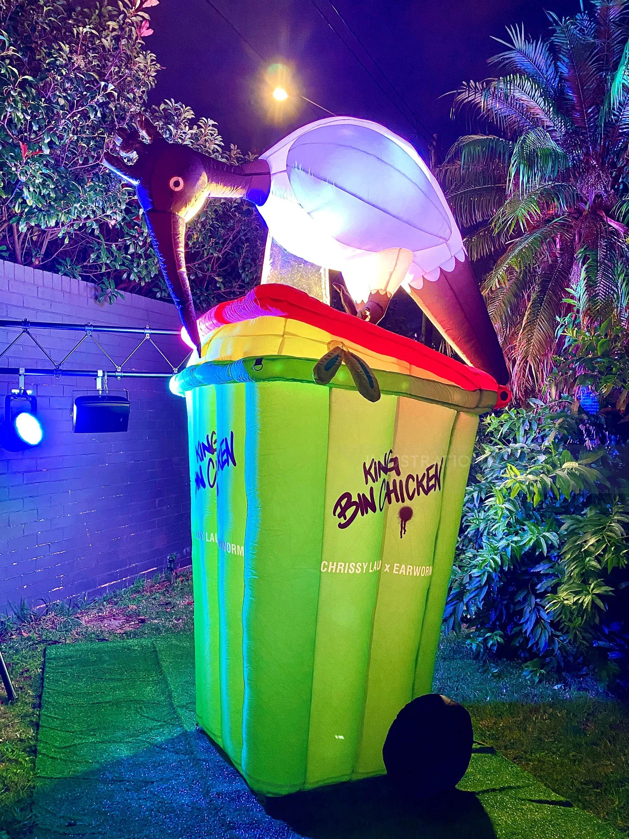 Brightly lit bird sculpture on green trash bin outdoors at night