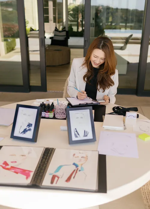 Woman drawing at table with sketches and art supplies by Elise Lee