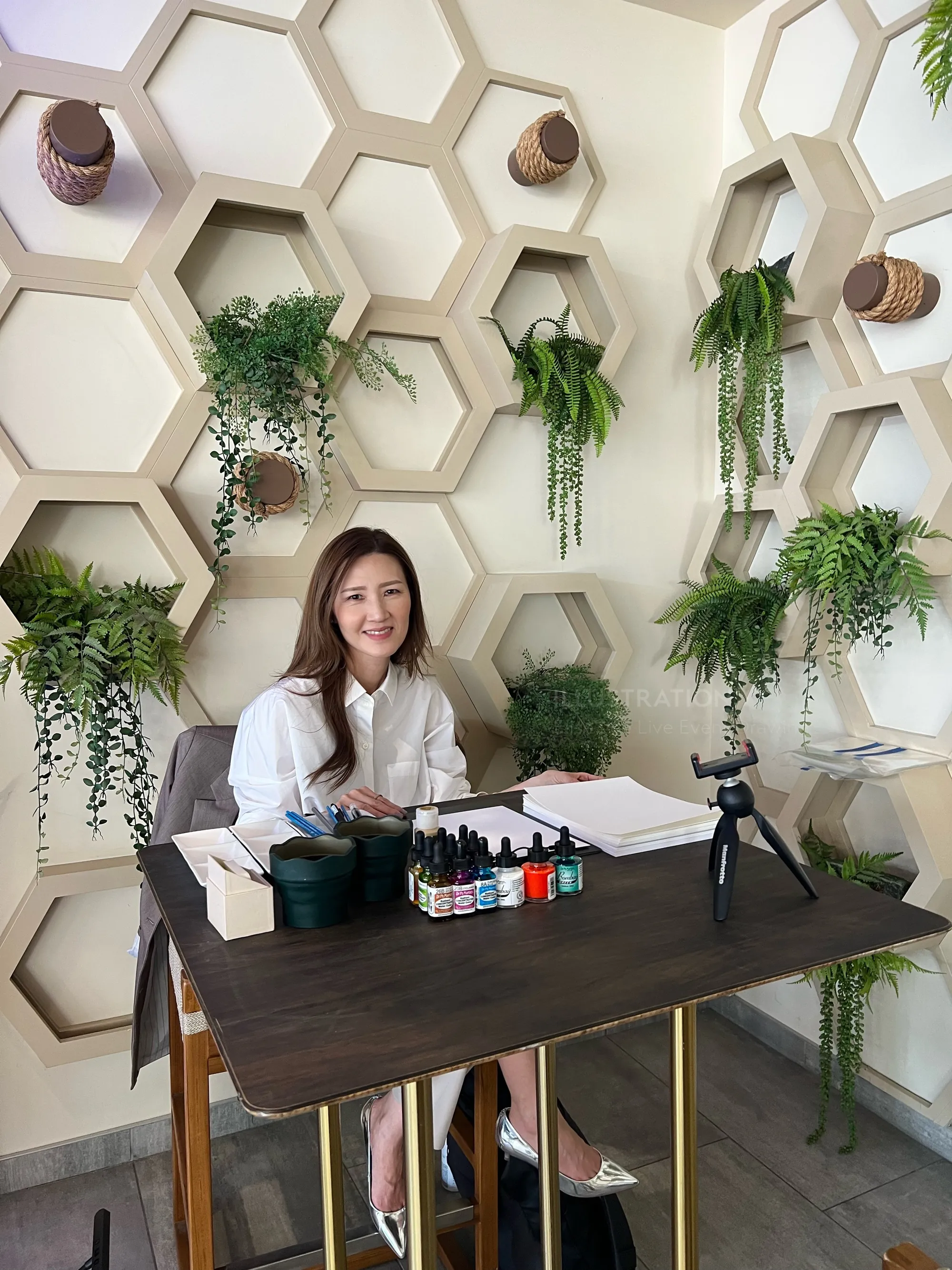 Woman sitting at table with art supplies and hexagonal plant wall by Elise Lee