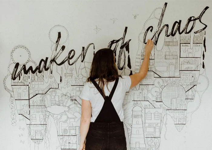 Woman writing the phrase maker of chaos on wall artwork by Ellie Compton