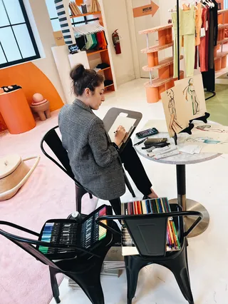 Woman sketching at table in bright studio with art supplies by Elly Azizian