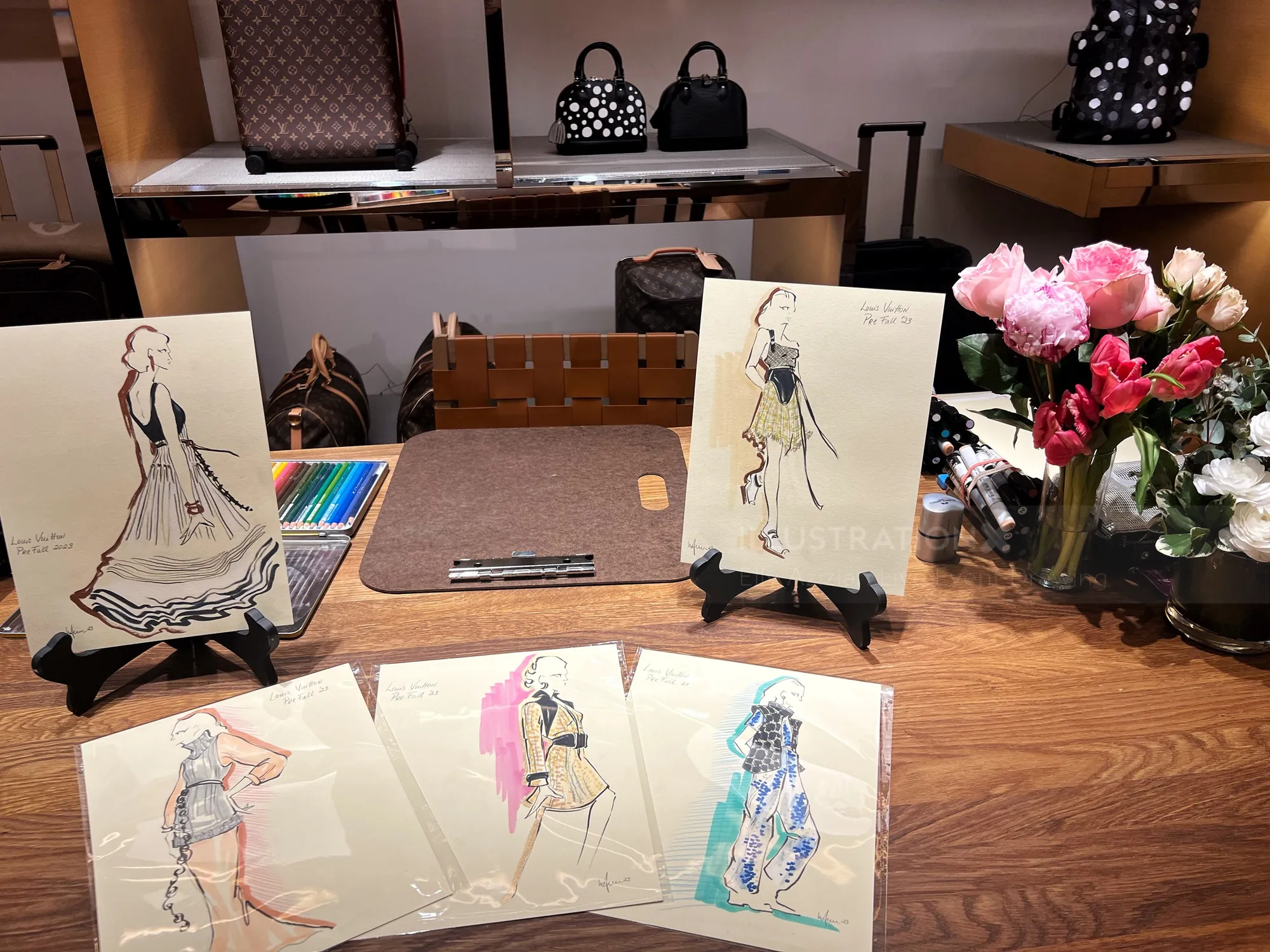 Bocetos de moda expuestos sobre una mesa con bolsos y flores por Elly Azizian