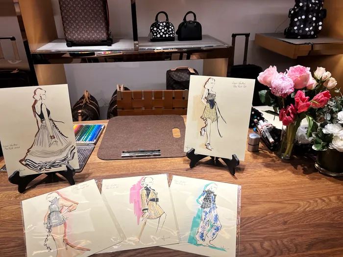 Fashion sketches displayed on table with bags and flowers by Elly Azizian