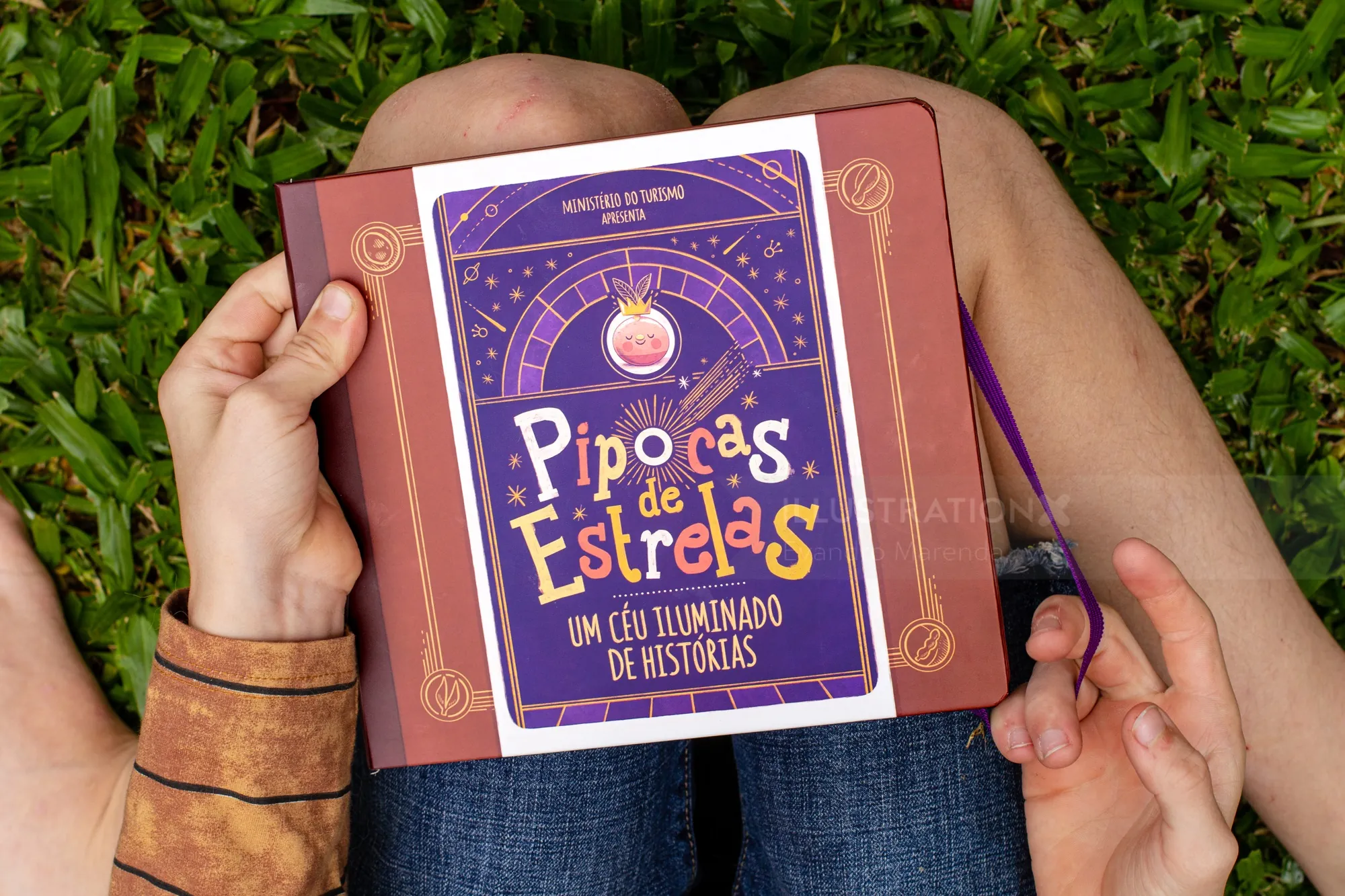 Hands holding open Pipocas de Estrelas book with grassy background