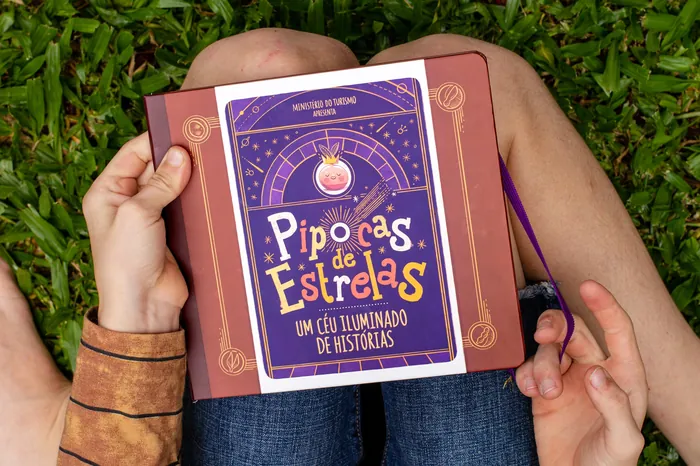 Hands holding open Pipocas de Estrelas book with grassy background
