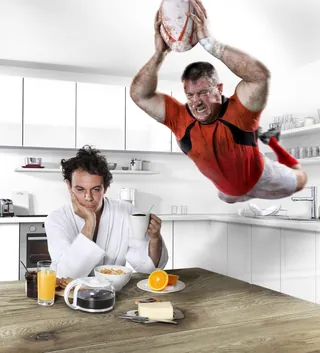 Rugby player diving mid-air over man eating breakfast in kitchen by Frank Neidhardt