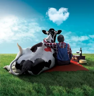 Man in overalls and cow sitting on grass with heart-shaped cloud by Frank Neidhardt