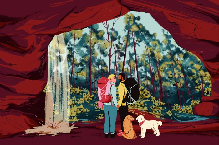 Two hikers with backpacks and dogs inside a cave near a waterfall.