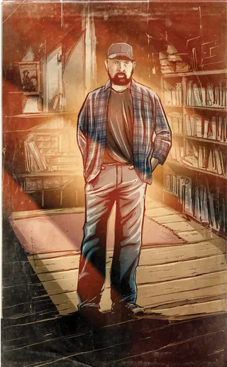 Bearded man wearing a cap and plaid shirt standing in a library.