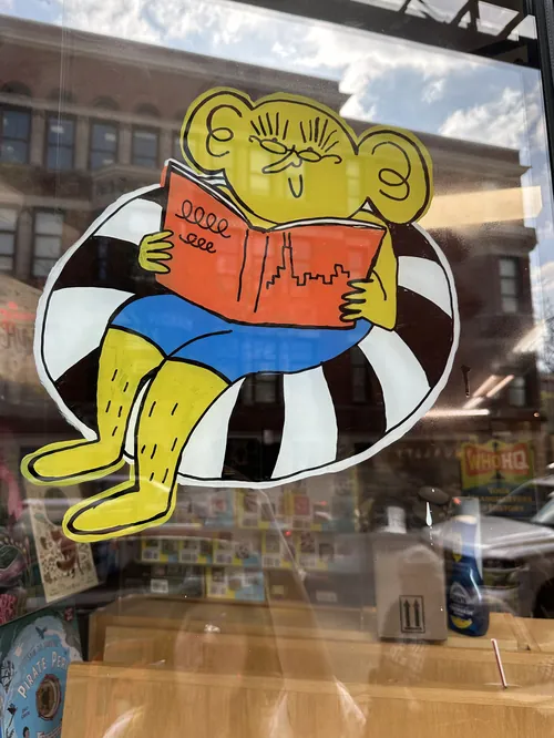 Personagem de desenho animado lendo um livro na vitrine de uma livraria, por Ollie Wootton.