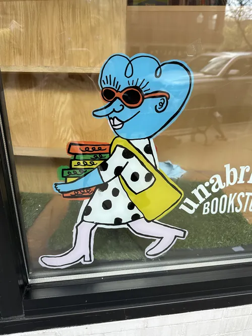 Mulher de desenho animado com cabelo azul carregando livros no mural "Livros Sem Cortes" de Ollie Wootton.