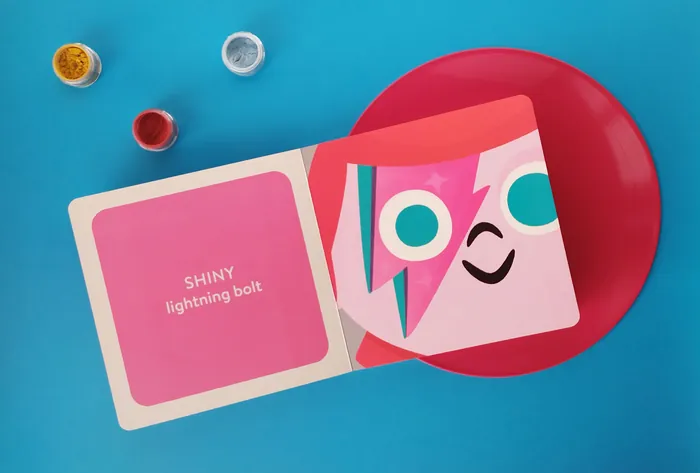 Open children's book with cartoon face and paint pots on blue background.