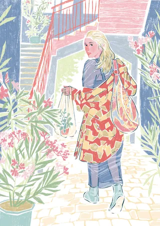 Woman in patterned coat walking among plants and flowers outdoors.