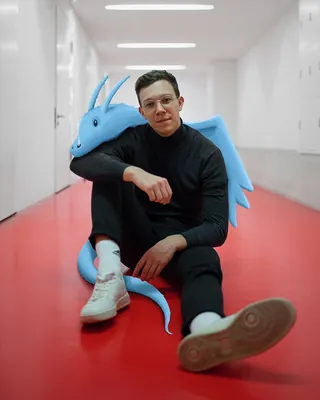 Man sitting beside a blue dragon sculpture in a white hallway by Samy Löwe