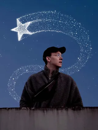 Man wearing glasses and cap looking up at glowing star trail in sky by Samy Löwe