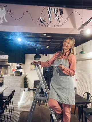 Woman in apron standing on ladder installing mural in cafe