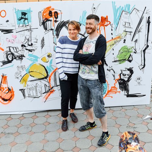 Two people standing in front of a large colorful mural canvas outdoors