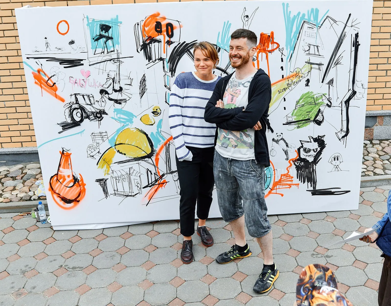 Two people standing in front of a large colorful mural canvas outdoors