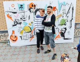 Two people standing in front of a large colorful mural canvas outdoors