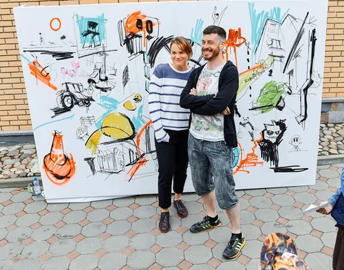 Two people standing in front of a large colorful mural canvas outdoors