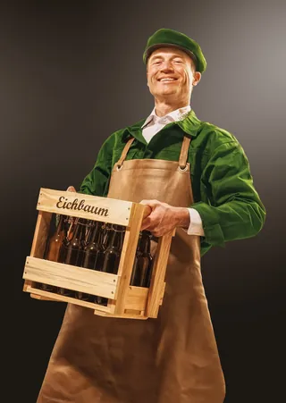 Smiling man in green shirt and brown apron holding Eichbaum beer crate by Tom Krieger