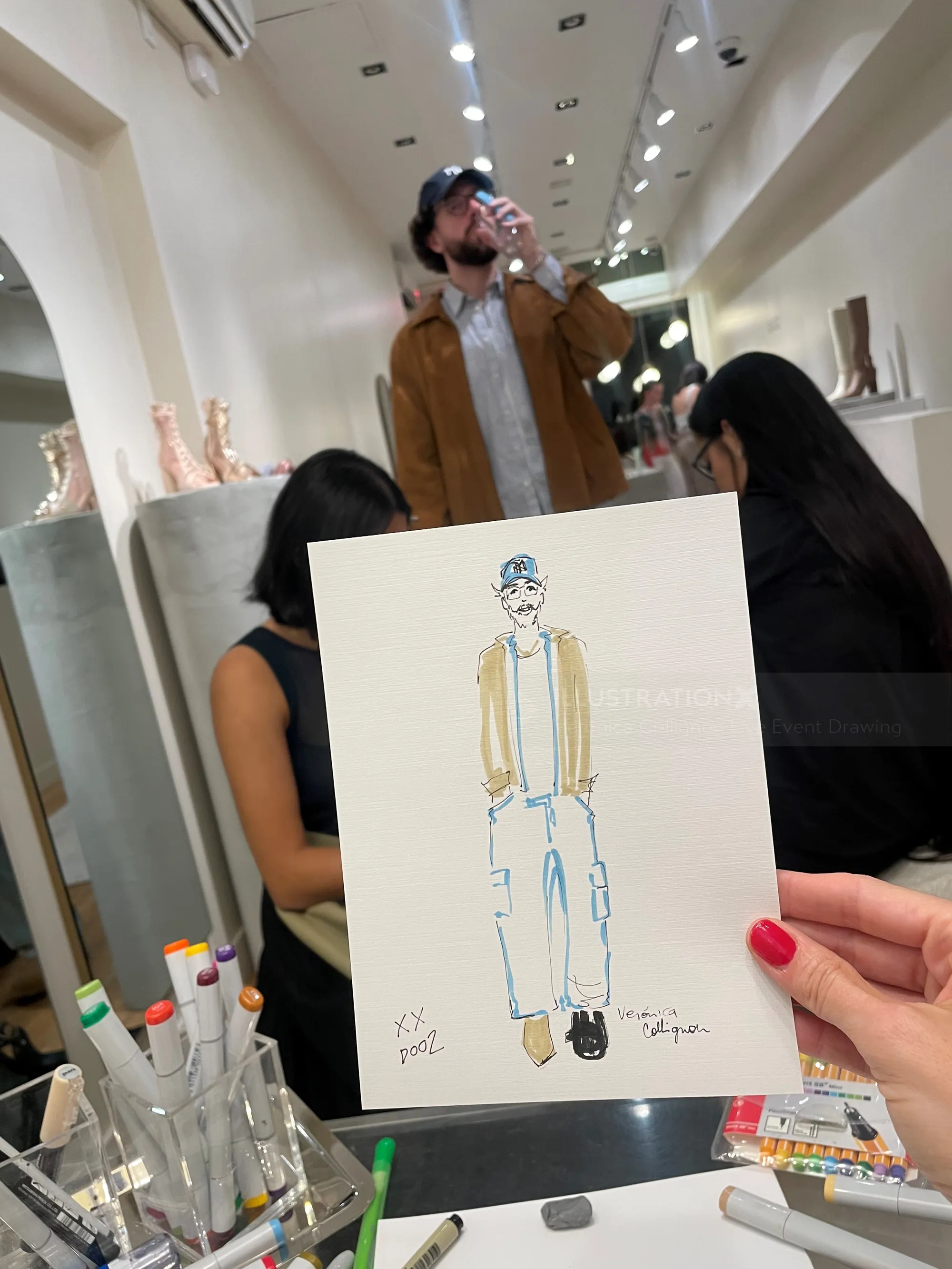 Eine Hand hält eine Modeskizze in einem Kunstatelier mit Models im Hintergrund.