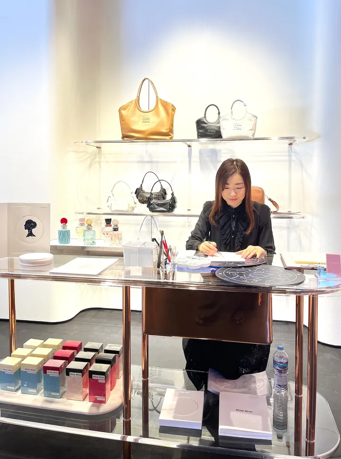 Woman writing at table with handbags displayed on shelves behind her
