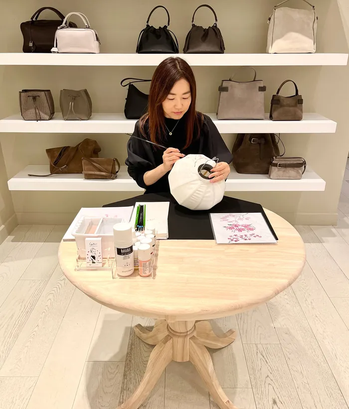Woman customizing white handbag at a table with handbags on shelves