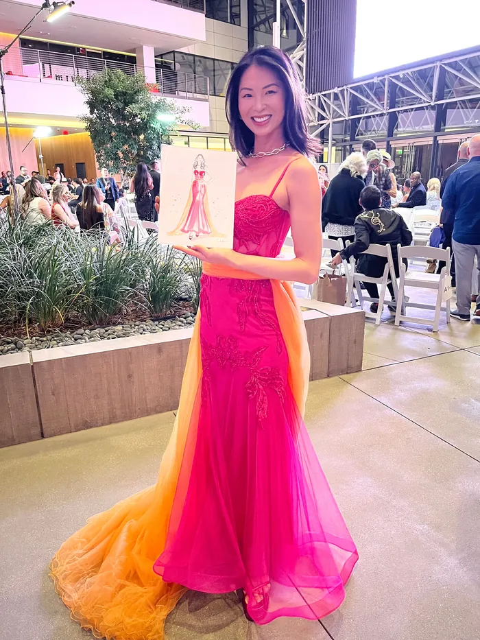 Woman in vibrant pink and orange gown holding a fashion sketch at event