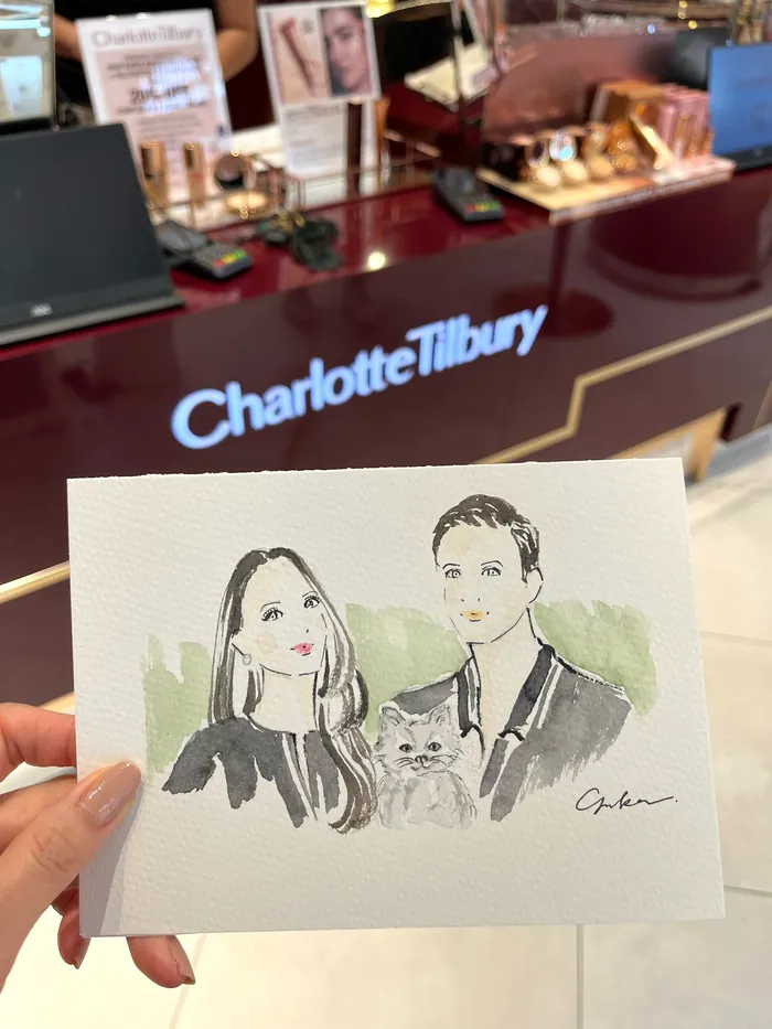 Hand holding a watercolor portrait of two women and a cat at a Charlotte Tilbury counter